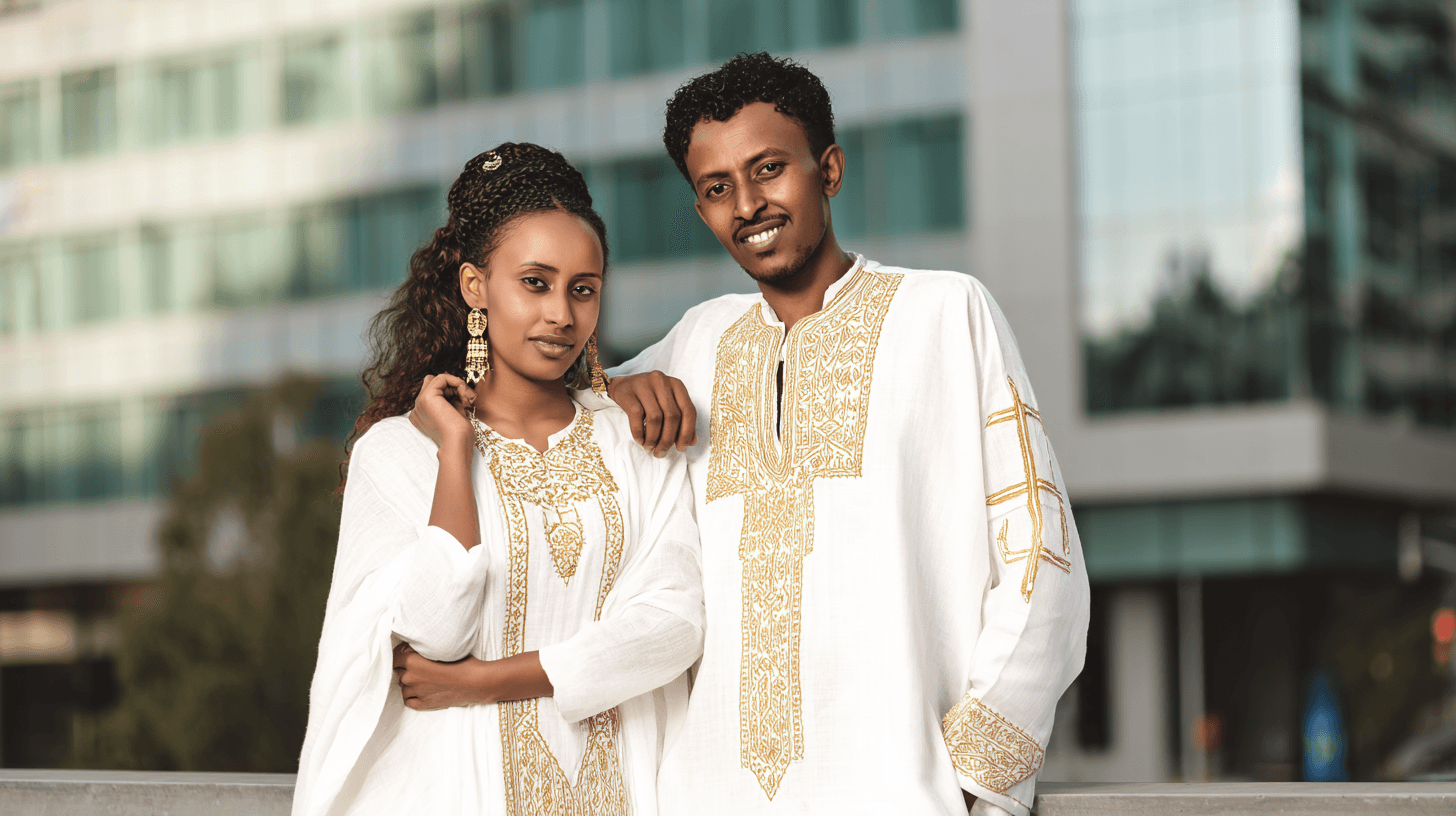 Ethiopian wedding couple standing outdoors in modern Addis Ababa wearing white habesha kemis with gold cross embroidery, glass building softly blurred behind