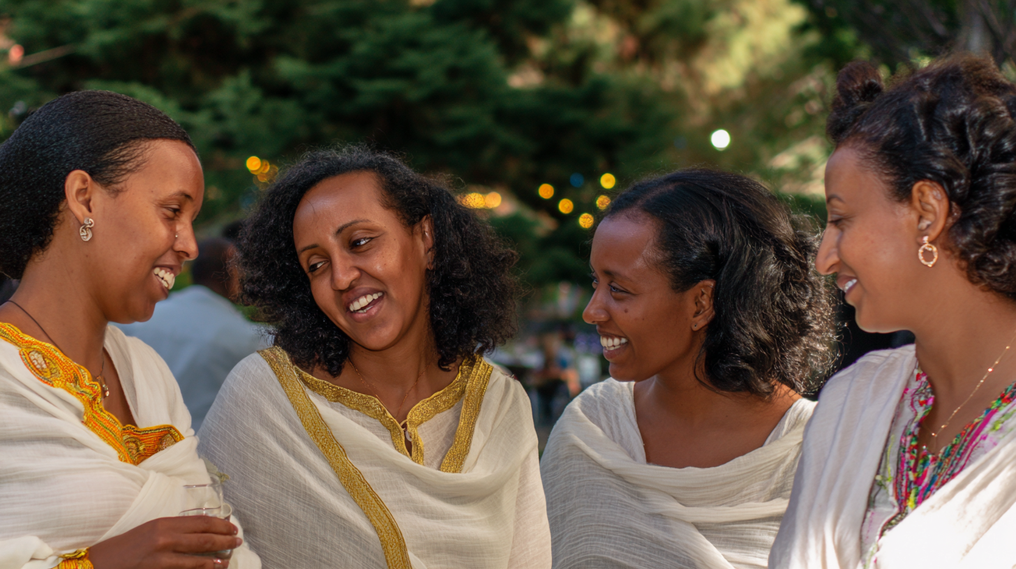 Four Ethiopian women wedding guests laughing at an outdoor reception, each wearing white habesha kemis in different styles with varying gold tibeb border details